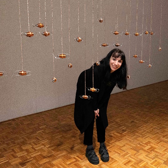 Artist Mimi Kind stands on parquetry wooden floor of the Chrissie cotter Gallery. She is smiling and wears all black, she has long hair and is bending down to interact with her bronze hanging sculptural artwork
